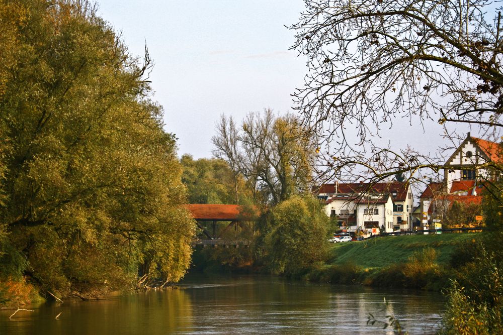 Ruhiges Wasser zwischen herbstlichen Bäumen, dahinter eine überdachte Holzbrücke mit rotem Dach und helle Häuser am Ufer