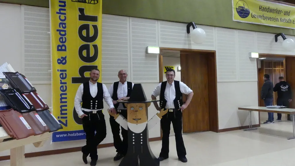 Drei Zimmerleute in Kluft mit einer Holzfigur vor dem Banner der Zimmerei